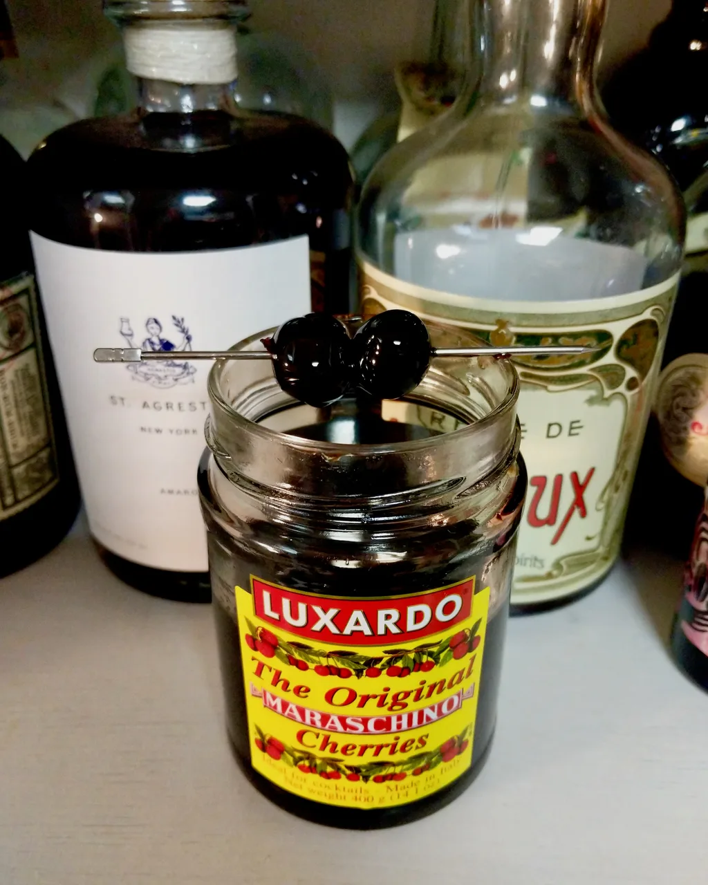 Cerejas marasca escuras em um espeto sobre o pote de cerejas Luxardo, com garrafas de licor ao fundo